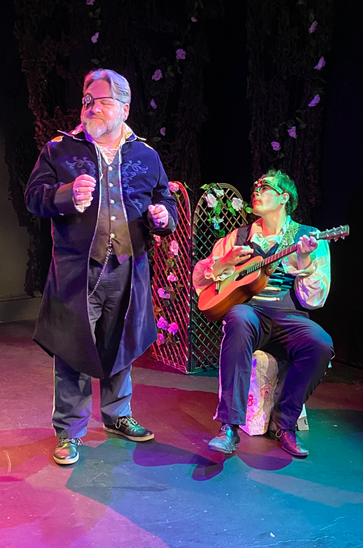 Don Pedro singing in the revels as Benedick plays guitar